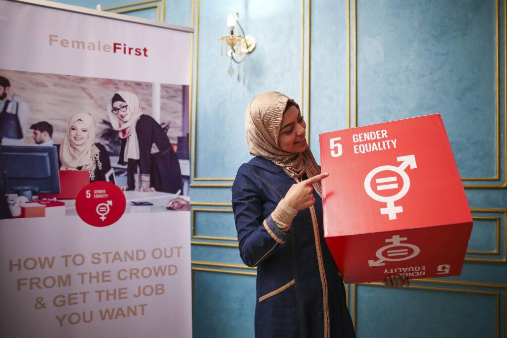 A woman in a hijab holding a red box with ‘gender equality’ written on it, next a poster for CTG’s Female First initiative, reading “How to stand out from the crowd & get the job you want”.